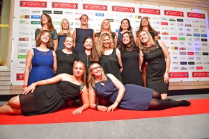 Mehrere Frauen posieren gemeinsam lächelnd auf einer Turnmatte vor einer Wand mit Sponsorenplakaten, für ein Gruppenfoto.