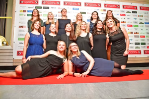 Gruppenporträt mehrerer Frauen in Abendkleidern vor einer Werbewand, lächelnd auf einer Turnmatte; Darstellung eines Teamfotos bei Veranstaltung.