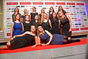 Porträt einer Gruppe fröhlicher Frauen in Abendkleidern auf rotem Teppich vor einer Sponsorwand, aufgenommen für ein Teamfoto.