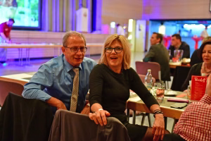Zwei Personen sitzen bei einer Veranstaltung und lächeln; sichtbares Tischgedeck und Konferenzkulisse im Hintergrund mit farbigen Lichtern.