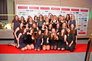 Gruppenfoto von mehreren Personen in dunkler Kleidung, nebeneinander vor einer Wand mit Sponsorenplaketten, posierend für eine Vereins-/Teamaufnahme.