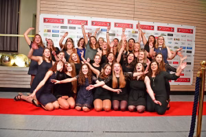 Gruppenfoto junger Frauen im Sportoutfit auf rotem Teppich; alle heben die Arme und jubeln vor einer Sponsorwand zur Feier eines sportlichen Erfolgs.