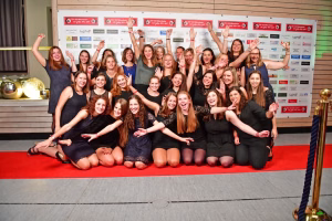 Gruppenfoto vieler junger Frauen auf rotem Teppich, alle winken und halten die Arme erhoben vor einer Wand mit Veranstaltungsplakaten.