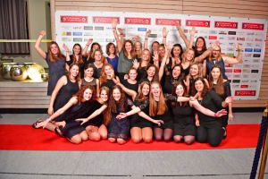 Gruppenfoto mehrerer Personen auf rotem Teppich; alle halten Arm- und Handzeichen hoch, wirken fröhlich vor einer Vereinswand als Gesamtaufnahme.
