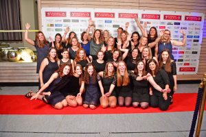 Gruppenfoto von Frauen und Mädchen in zwei Reihen auf rotem Teppich, lächelnd und winkend, vor einer Sponsorwand bei einer Veranstaltung.