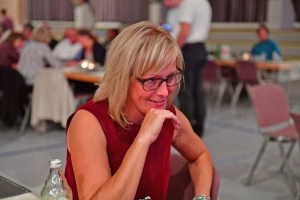 Lächelnde Frau mit Brille und rotem Top sitzt am Tisch und stützt den Kopf auf die Hand, im Hintergrund eine festliche Tischrunde.