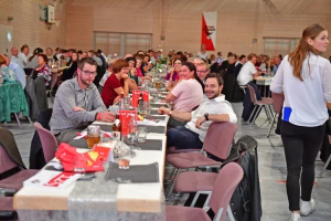 Gedeckte Tafeln in einer Sporthalle: Menschen sitzen und essen, im Vordergrund steht eine Serviette mit Getränke- und Platzkärtchen.
