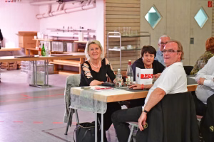 Mehrere Personen sitzen an einem gedeckten Tisch in einer Halle; am Tisch stehen Getränke und Unterlagen, im Vordergrund unterhalten sie sich.