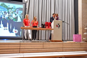 Podium einer Veranstaltung: drei Personen in roten Shirts und Anzug hinter einem Rednerpult, im Hintergrund ein Musikerensemble auf Leinwand.
