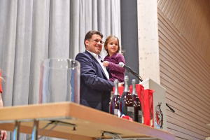 Mann und Kind am Rednerpult mit Mikrofon; vor ihnen Getränkeflaschen und Vorhänge, im Hintergrund Holzpaneele.