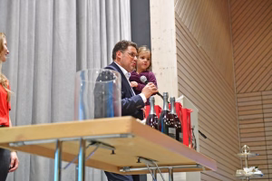 Mann mit Kind hält am Mikrofon eine Rede; Tische mit Getränkeflaschen im Vordergrund vor grauen Vorhängen und Holzdecke.