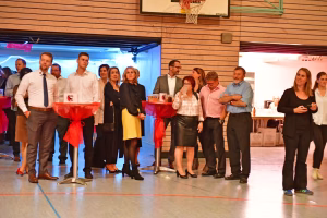 Gruppe von Menschen in einem Raum mit roter Deko am Tisch, einige stehen im Halbrund und wirken wie bei einer Veranstaltung oder Rede.