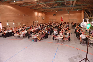 Großer Saal mit mehreren mit gedeckten Tischen sitzenden Gästen und Bühne; Veranstaltung mit Scheinwerfern und Leinwänden.