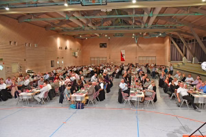 Überfüllte Sporthalle mit langen Tischen, Speisen und Getränken; viele Teilnehmende sitzen und stehen bei einer gemeinsamen Veranstaltung.