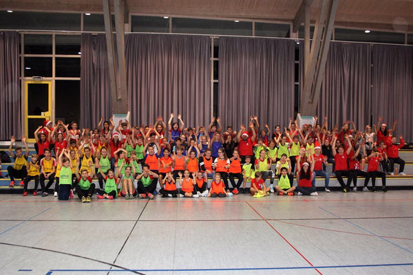 Gruppenfoto vieler Kinder und Betreuer in Sporthalle mit einheitlichen farbigen T-Shirts, gemeinsam winkend zur Feier eines Anlasses.