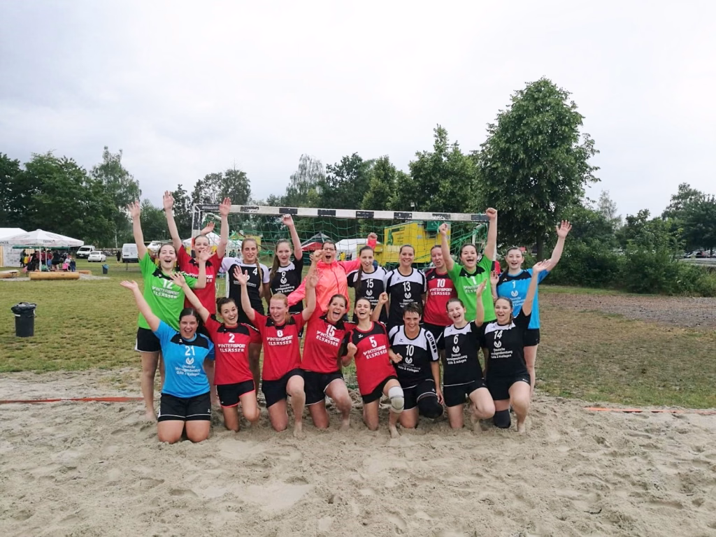 Mehrere Beachvolleyballspielerinnen jubeln im Sand, bilden in zwei Reihen ein Teamfoto und halten die Hände hoch vor einem Tornetz.