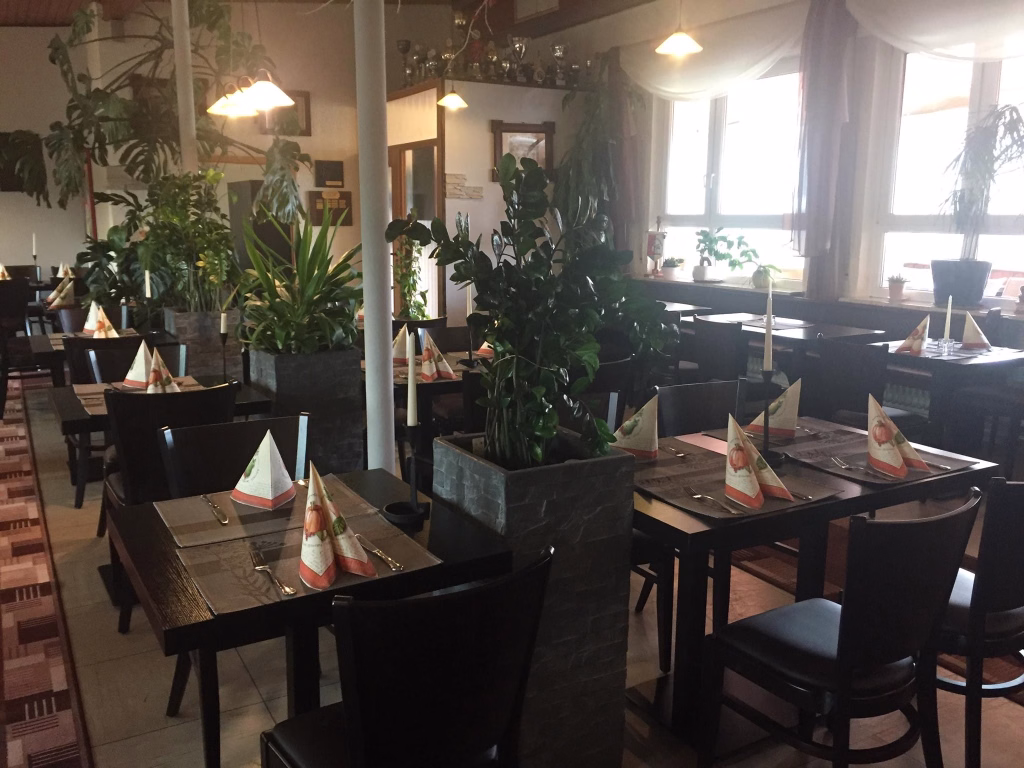 Restaurantinnenraum mit gedeckten Tischen, Kerzen und mehreren Topfpflanzen; warme Beleuchtung und Fensterlicht schaffen eine einladende Atmosphäre.