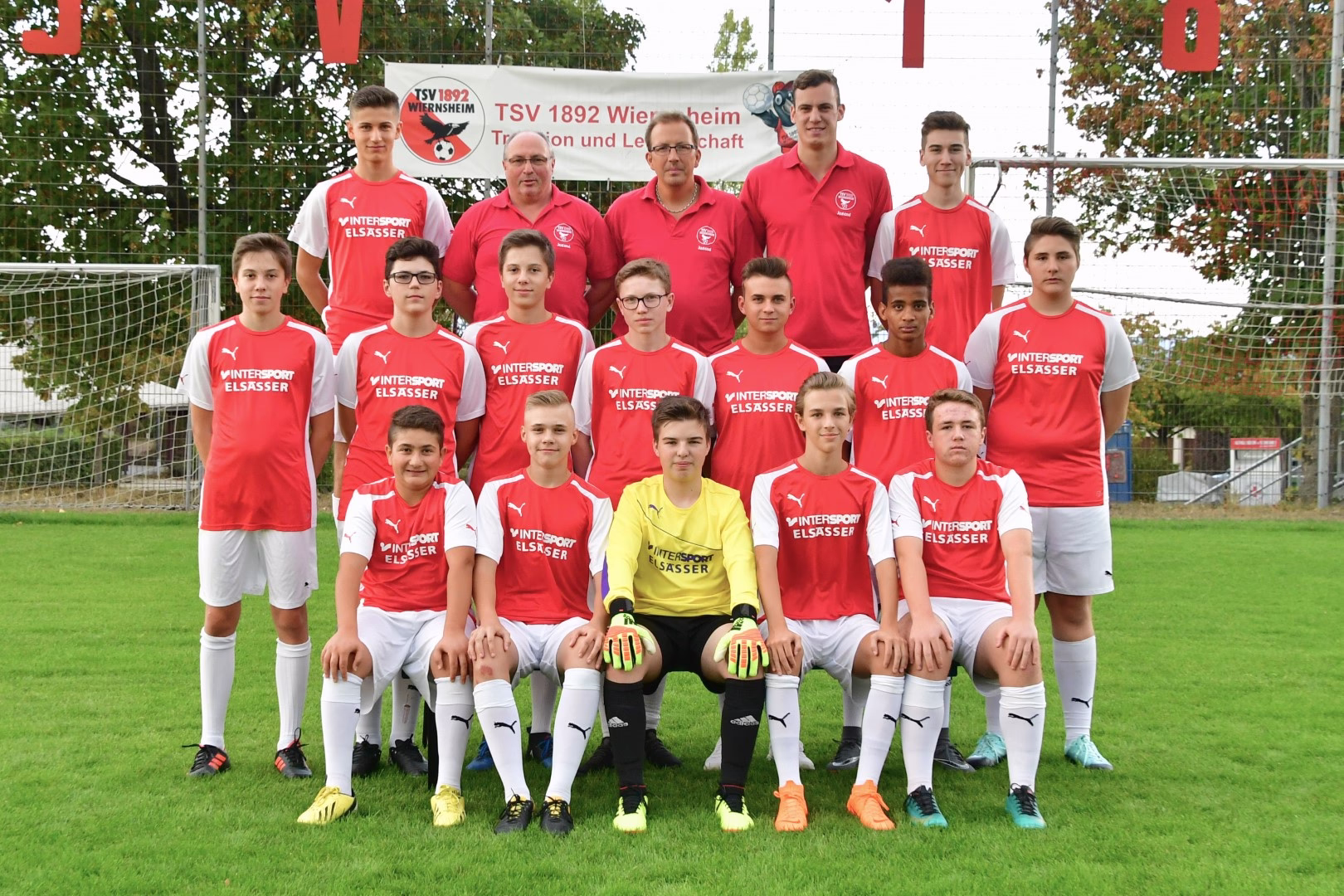 Mannschaftsaufstellung einer Jugend-Fußballmannschaft im Stadion: mehrere Spieler in roten/weißen Trikots posieren vor dem Tor, zentral ein gelbes Torwarttrikot.