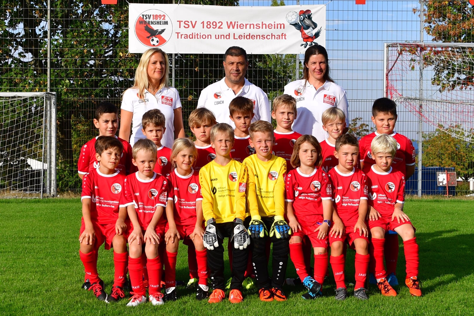 Fußballmannschaft mit Trainerteam in Rot und Gelb vor dem Tor, Banner im Hintergrund; Gruppenfoto für die Teampräsentation.