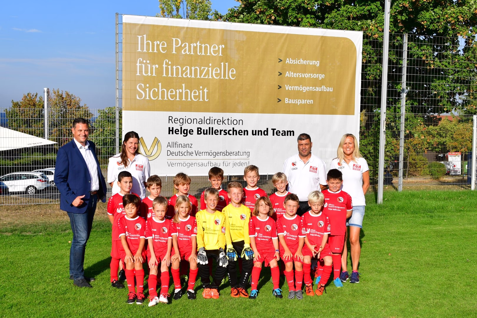 Mannschaftsbild der F-Jugend auf Rasen vor Werbebanner; Trainer und Kinder stehen für Sponsoring/Unterstützung sichtbar nebeneinander.