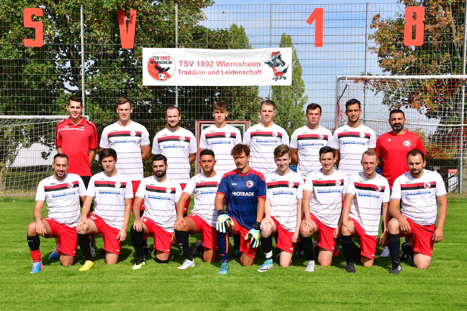 Fußballteam-Foto: Spieler in zwei Reihen auf dem Rasen vor Tor und Zaun, Banner mit großen Zahlen zur Einordnung des Teams/Events.