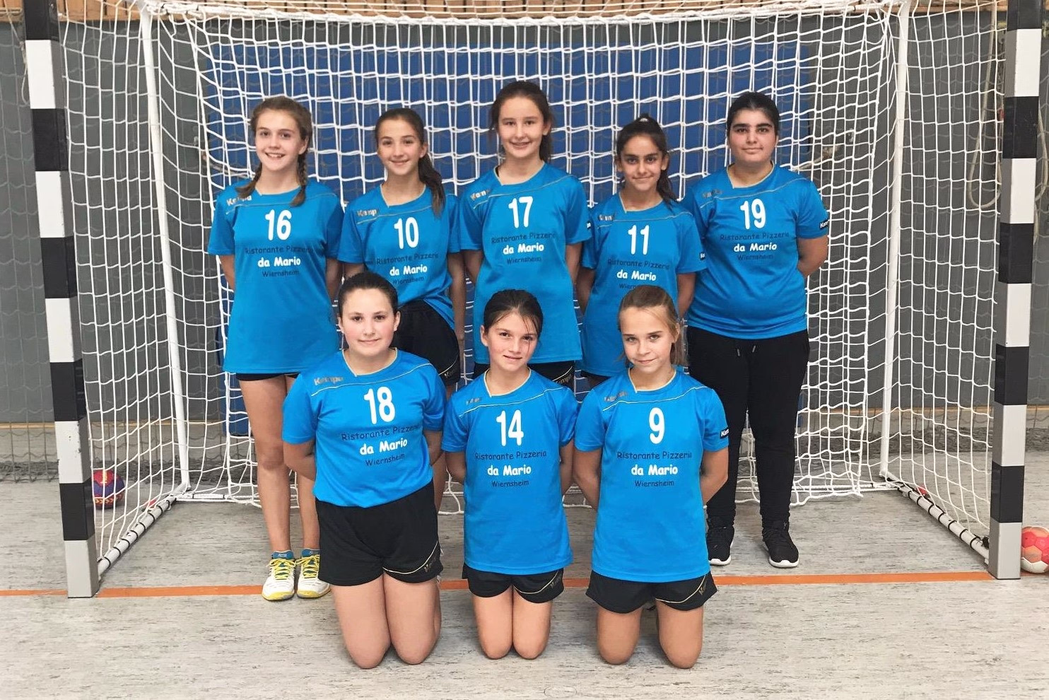 Mannschaftsbild von acht Mädchen in blauen Sportshirts vor einem Fußballtor; Trikotnummern 9,10,11,14,16,17,18,19 sichtbar.