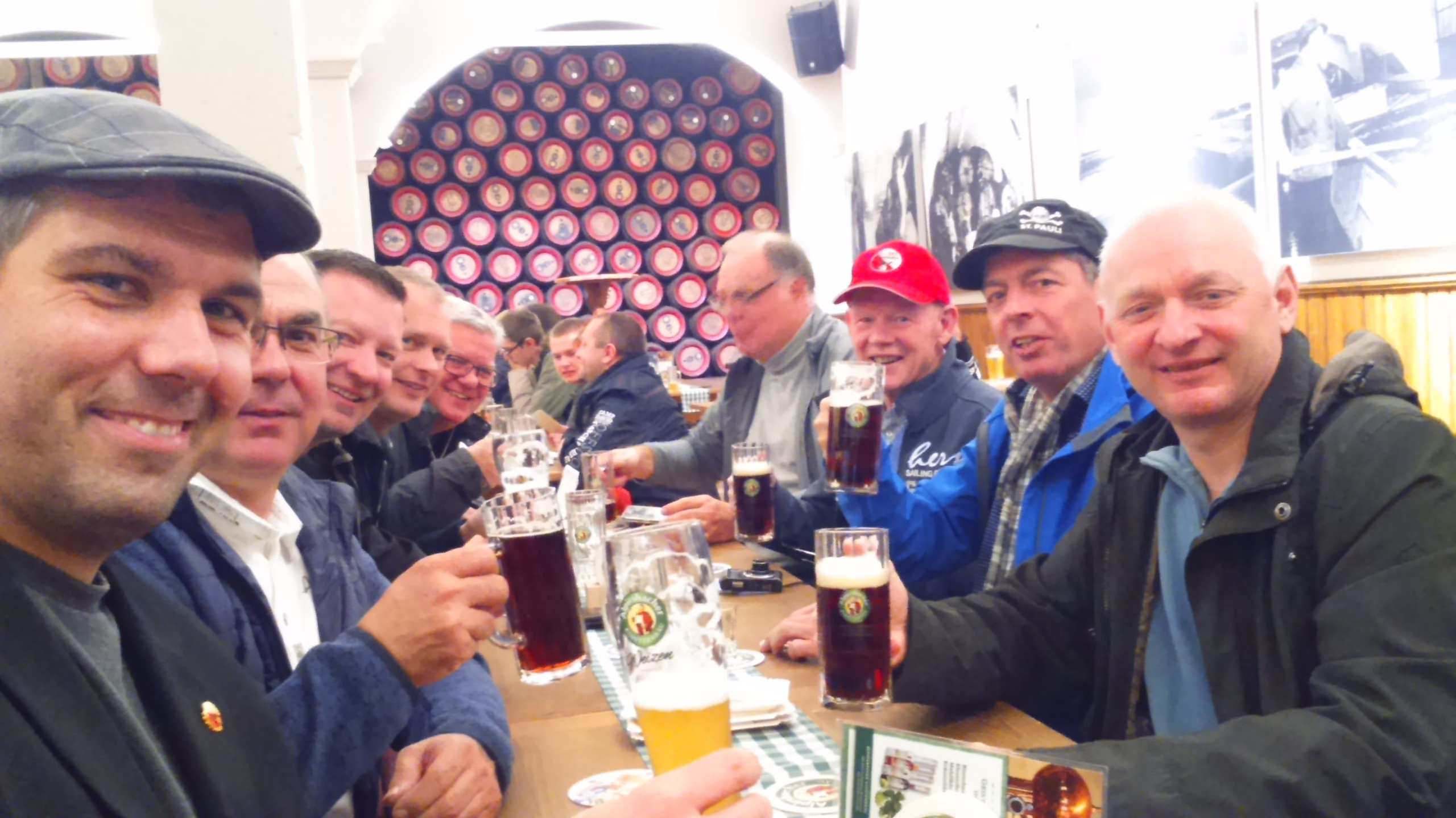 Mehrere Männer sitzen an einem Tisch, stoßen mit Biergläsern an; Hintergrund mit dekorativem Kreis-Muster, Innenbereich einer Veranstaltung.