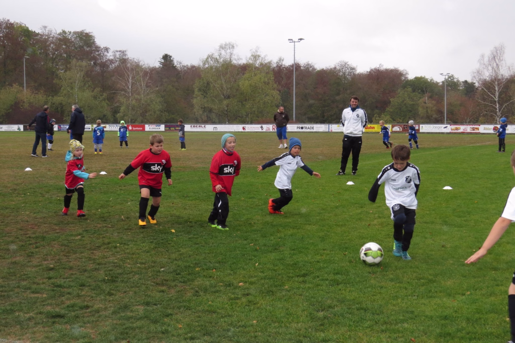 Jugendliche spielen auf einem Fußballfeld; mehrere Kinder laufen und schießen nach dem Ball, im Hintergrund Zuschauer und Vereinsbanner.