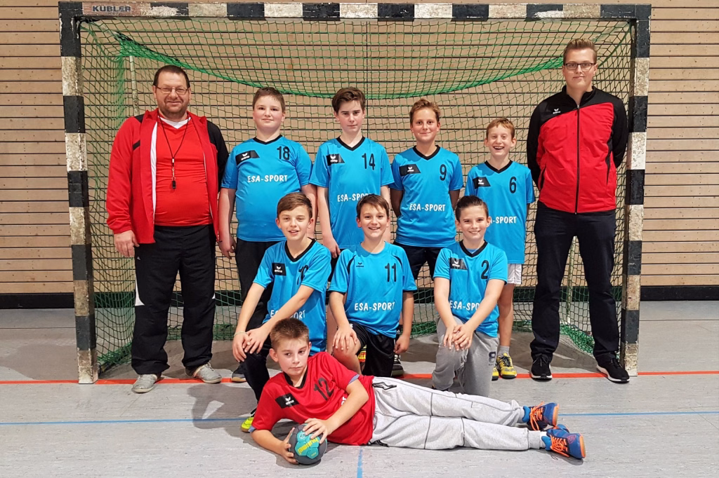 Mannschaftsfoto mit Trainer und mehreren Handballspielern vor einem Tor, im Vordergrund ein liegender Spieler—zur Teamdarstellung.