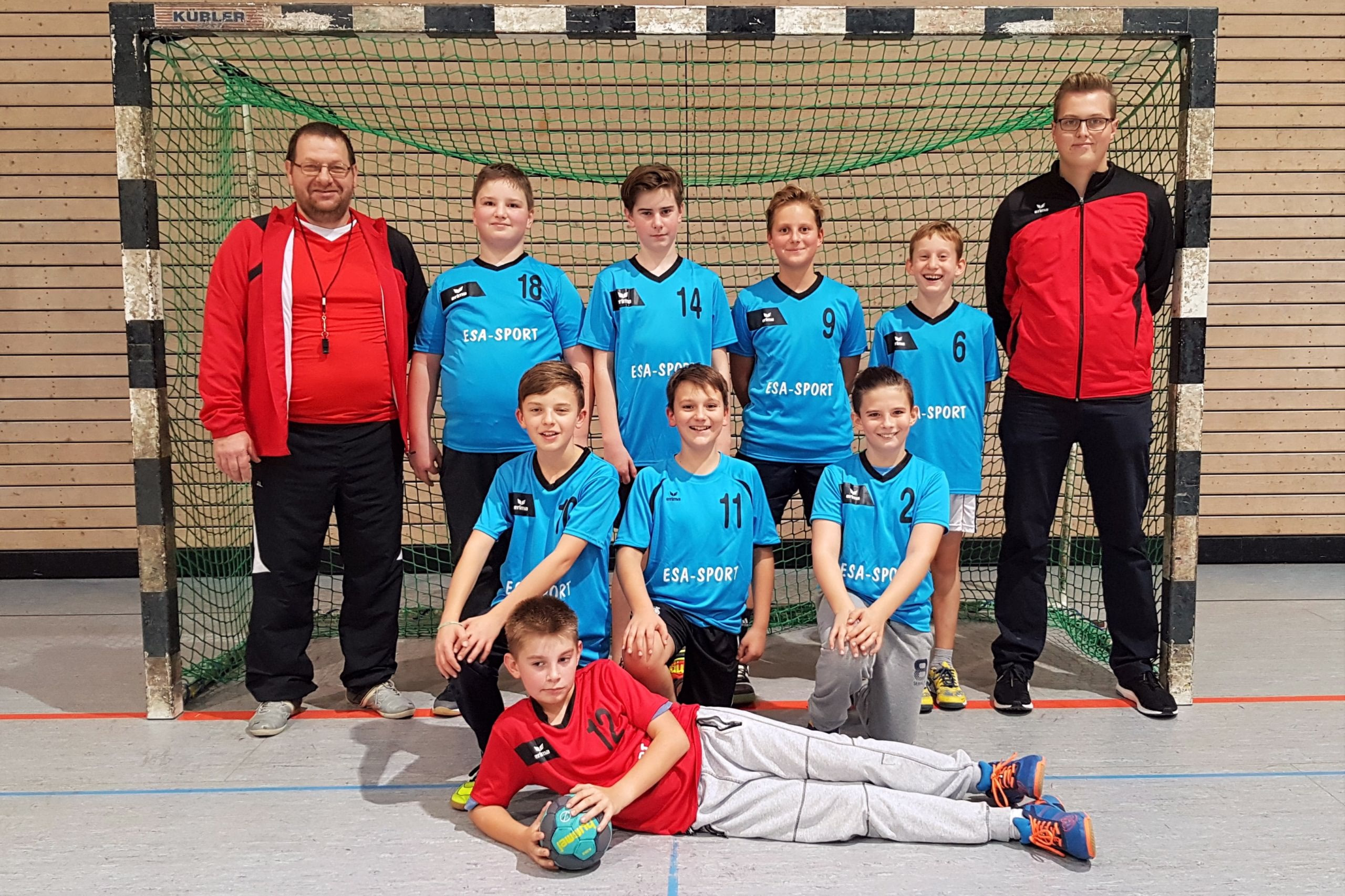 Mannschaftsfoto mit Trainer und mehreren Handballspielern vor einem Tor, im Vordergrund ein liegender Spieler—zur Teamdarstellung.