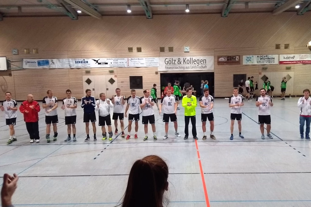 Handballmannschaft steht vor Zuschauern in einer Sporthalle, trägt Trikots in Weiß, Grün und Blau, in Vorbereitung zur Begrüßung.