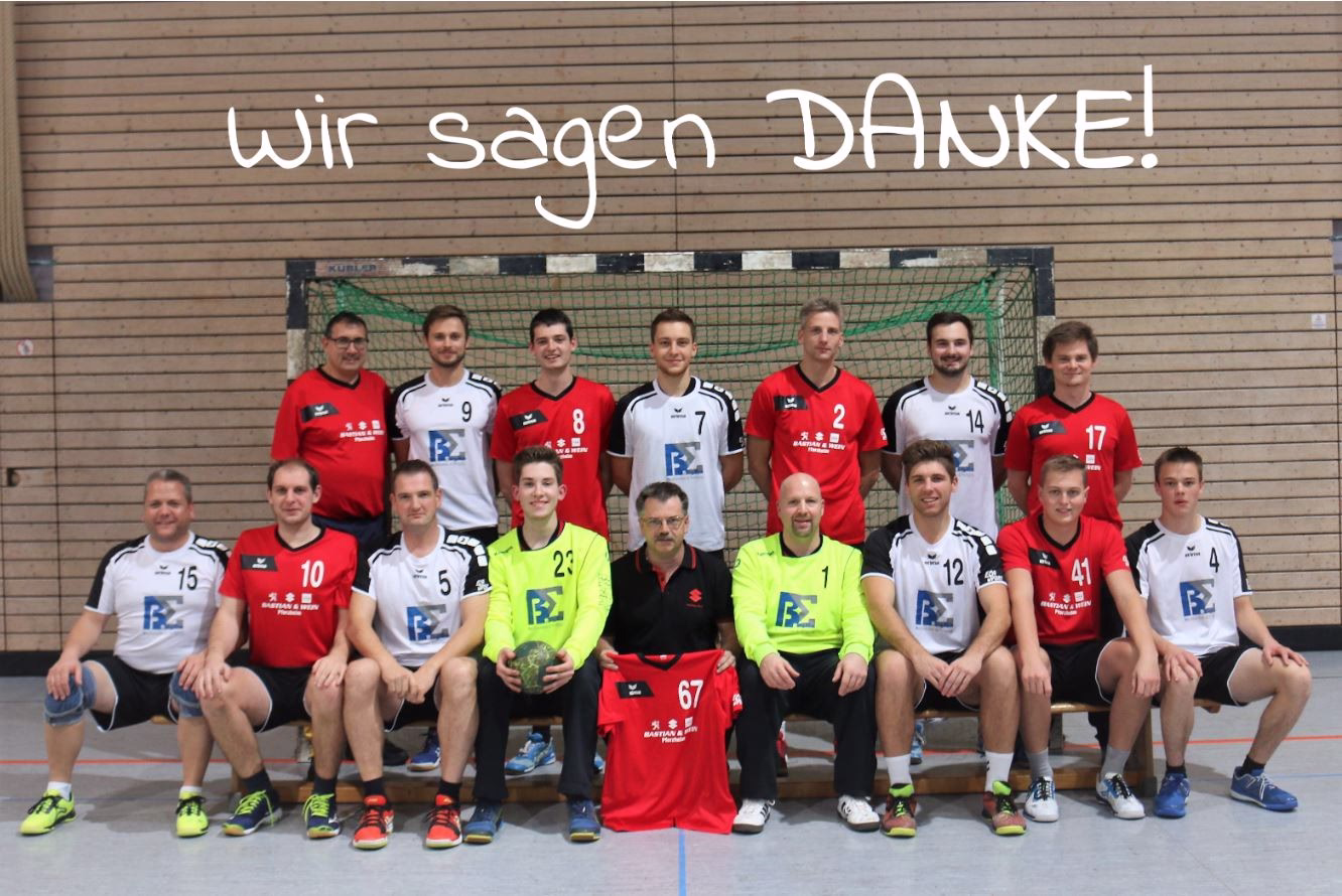 Gruppenfoto einer Handballmannschaft vor Hallenwand mit großen Schriftzug „Wir sagen DANKE!“ als Dankesbotschaft.