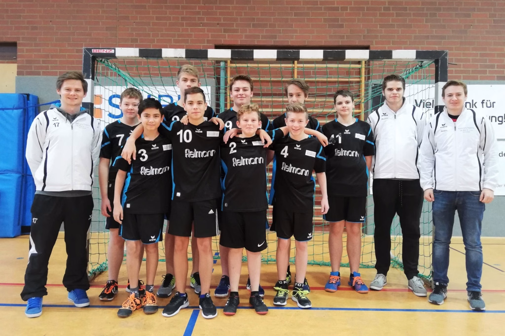 Sportteam in der Halle posiert vor einem Handballtor, trägt Trikots und steht gemeinsam für ein Mannschaftsfoto.