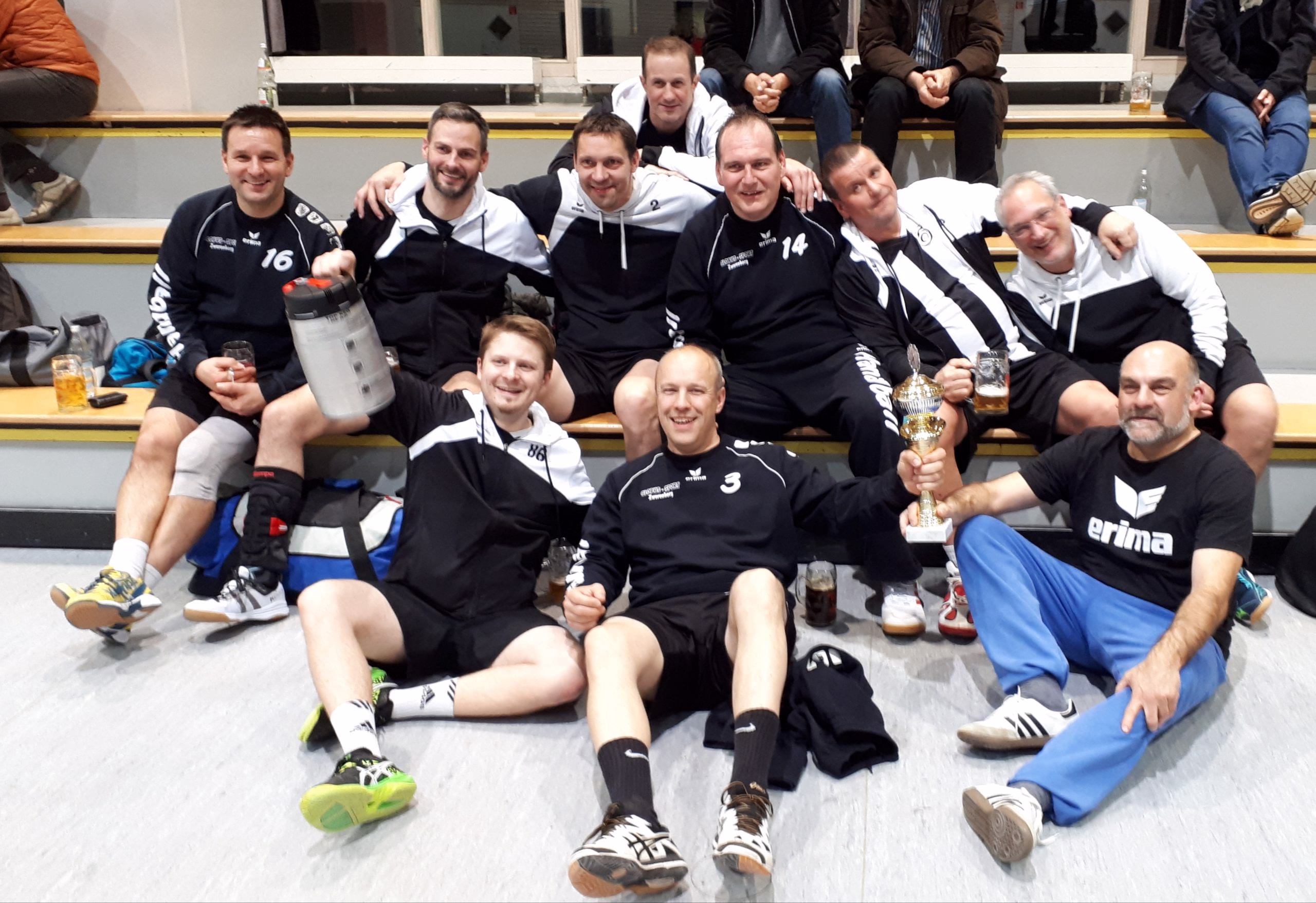 Mannschaft feiert gemeinsam im Innenraum: mehrere Personen sitzen eng auf einer Bank, halten Pokal und Drinks, einige lachen und posieren.