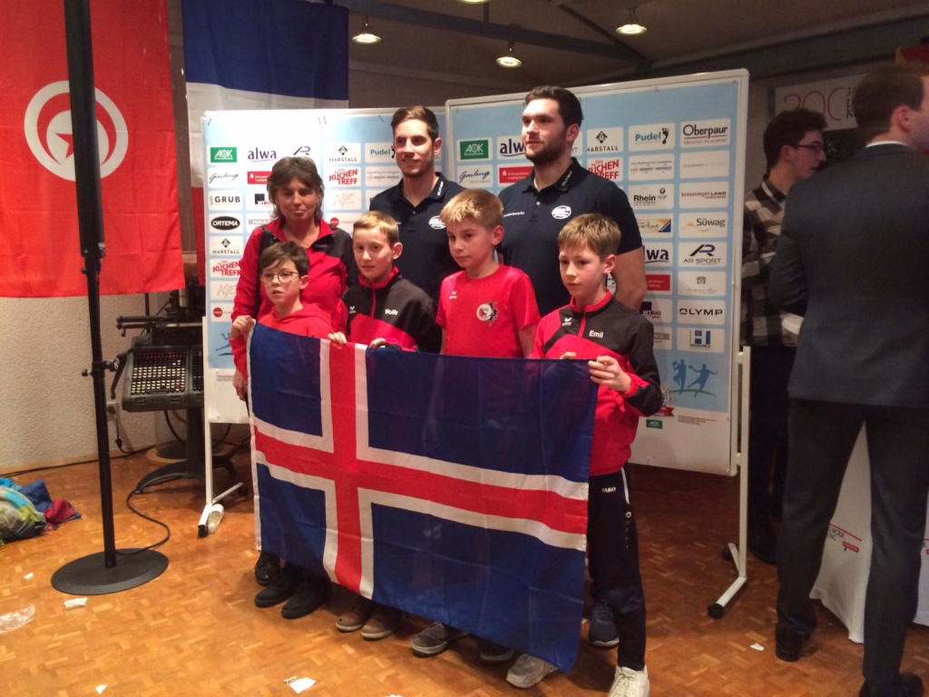E-Jugend tritt als Island bei der Mini-WM an 7 Gruppe aus Erwachsenen und Kindern posiert in Sportkleidung vor einer Sponsorwand, hält gemeinsam die Flagge eines Landes als Mannschaftsfoto.