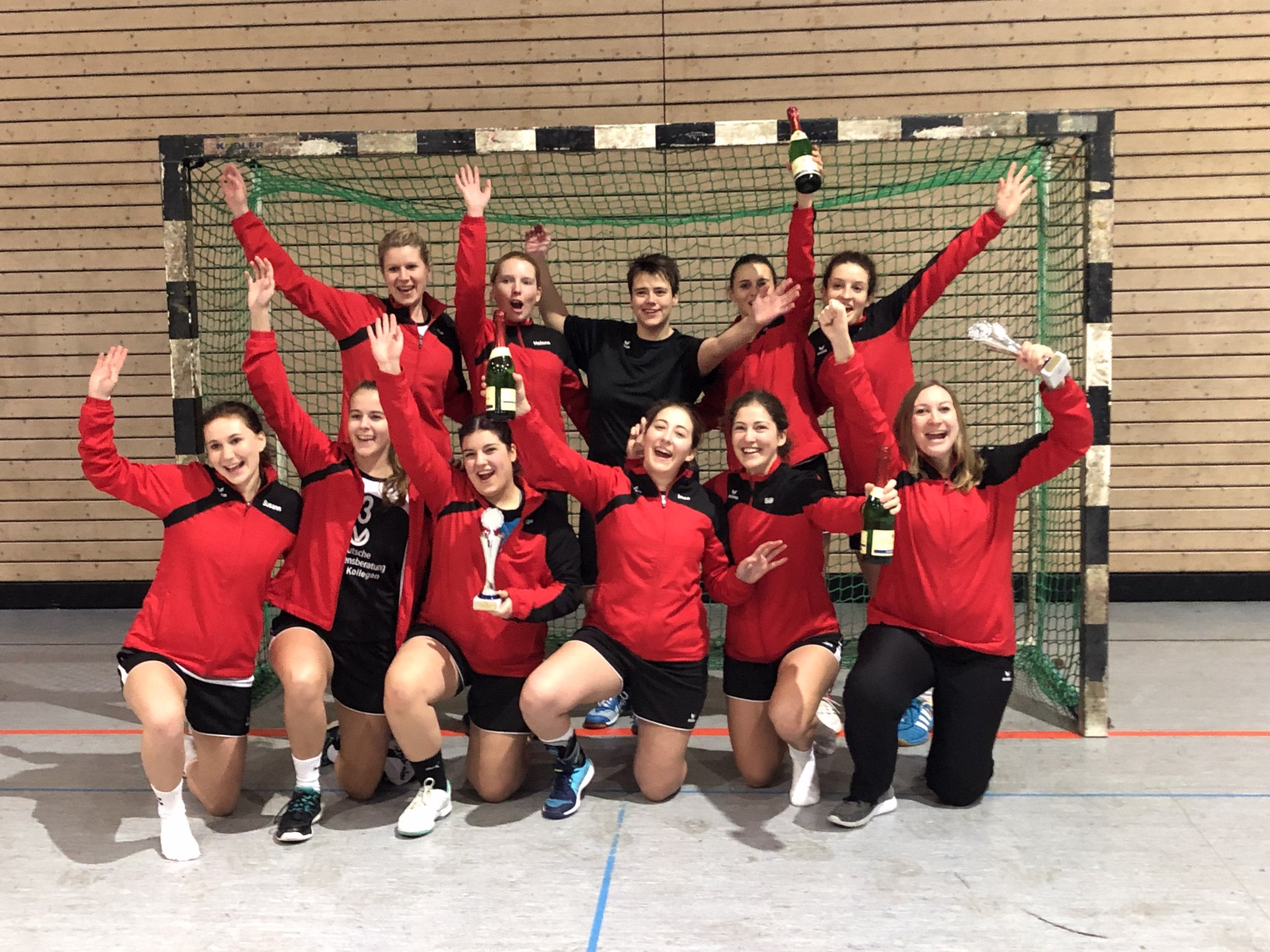 Mannschaft in Sportkleidung posiert vor einem Handballtor und streckt die Hände nach oben; Trophäe und Siegergruß im Turnierkontext.