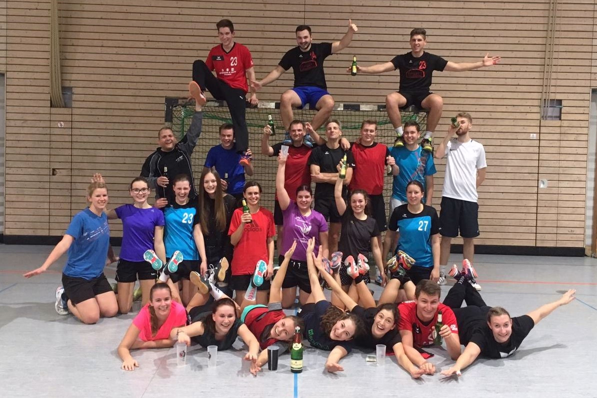 Gruppe trainierender Personen in einer Sporthalle posiert lachend auf dem Boden und steht; einige halten Sektflaschen als Siegerfoto.