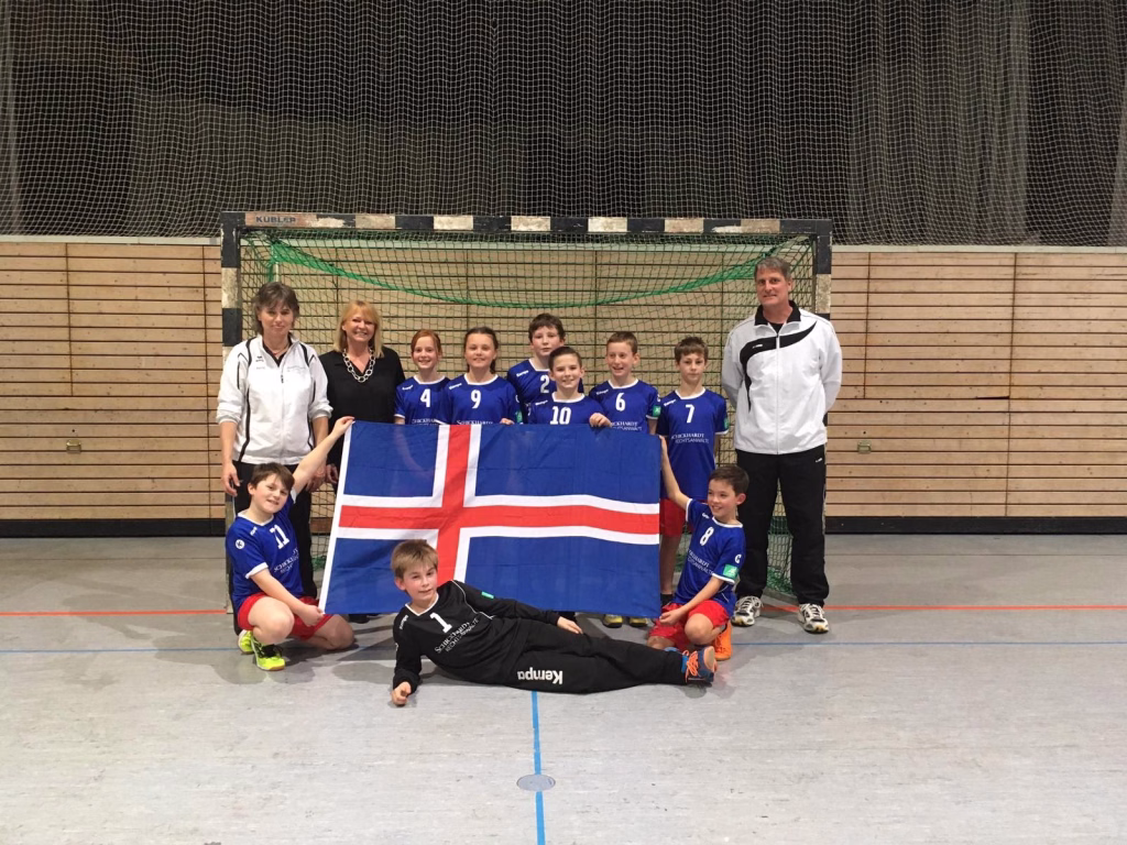 Mini-Island nimmt seine WM-Trikots in Empfang 3 Sportteam mit Kindern und zwei Erwachsenen in Hallenkleidung vor dem Tor, hält gemeinsam eine große Flagge.