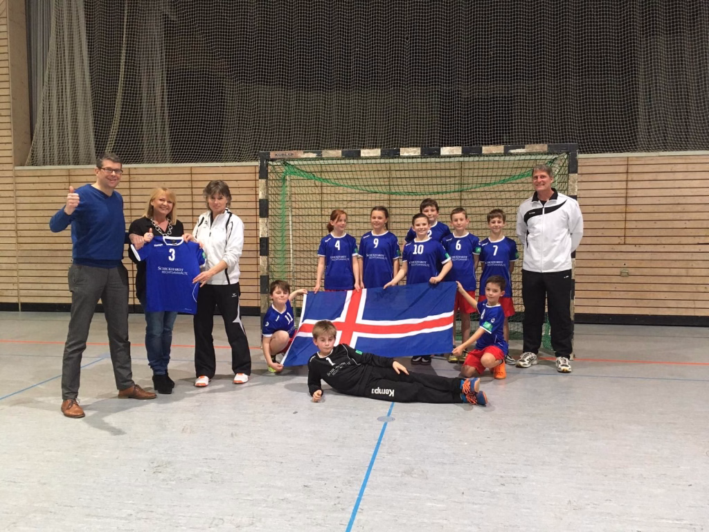 Mini-Island nimmt seine WM-Trikots in Empfang 8 Sportteam mit Trainer vor einem Handballtor in der Halle; Kinder posieren mit Trikots und Flagge für ein Mannschaftsfoto.