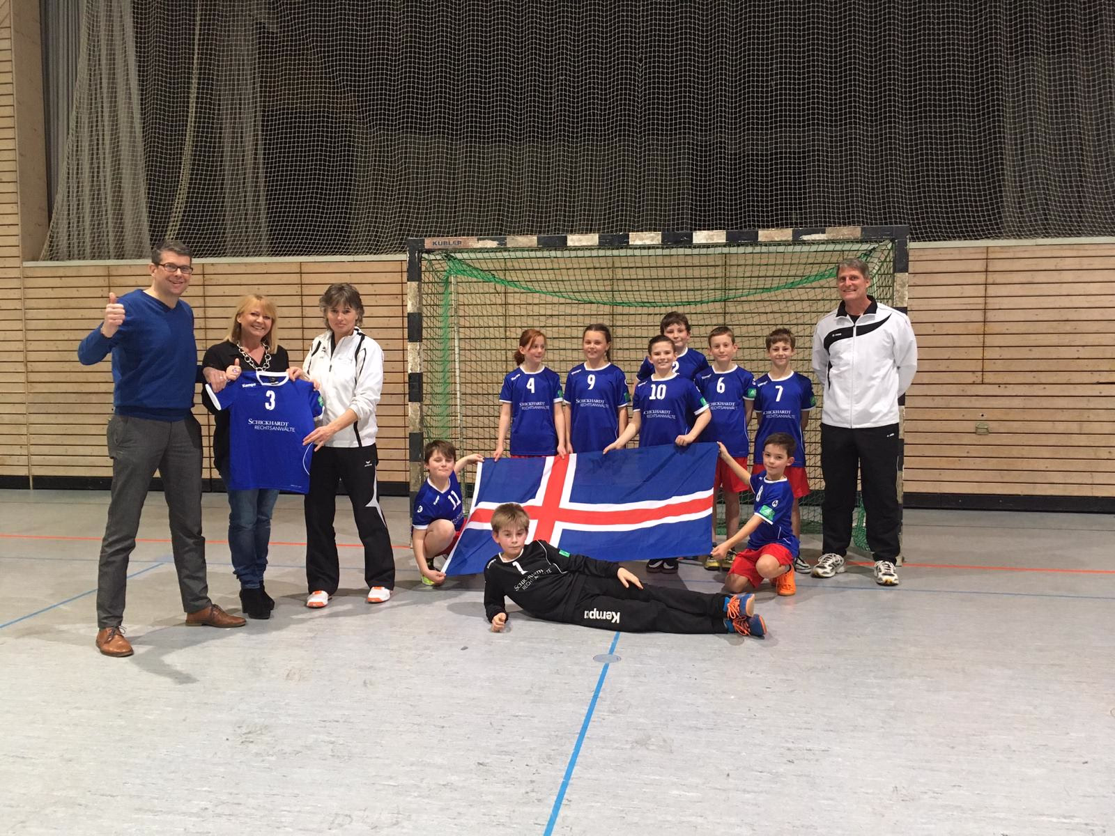 Sportteam mit Trainer vor einem Handballtor in der Halle; Kinder posieren mit Trikots und Flagge für ein Mannschaftsfoto.