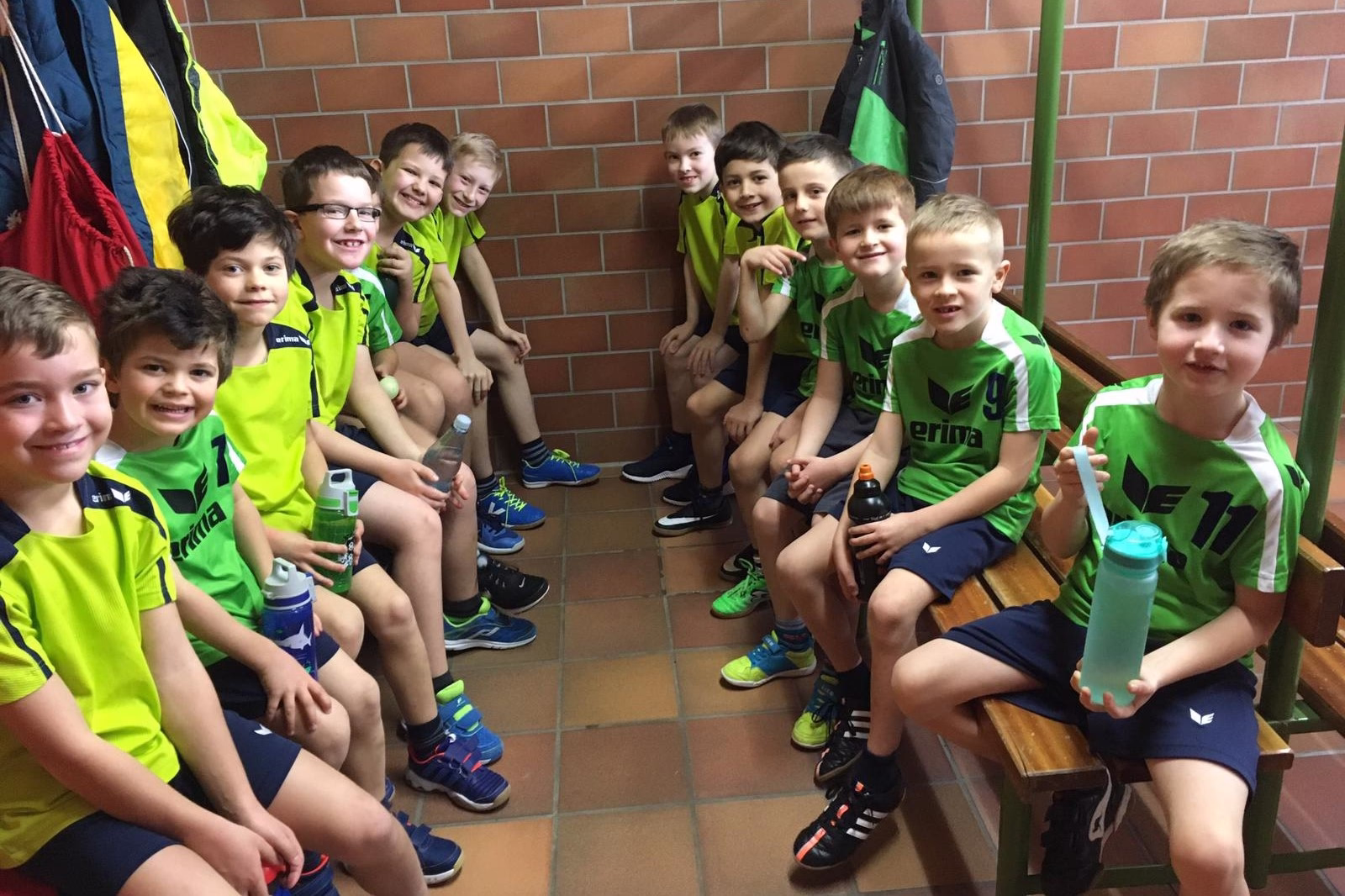 Gruppe von Kindern im Sportdress sitzt auf einer Treppenstufe an der Backsteinwand, einige halten Getränkeflaschen und lächeln.