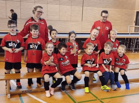 Gruppe von Kindern und zwei Erwachsenen in Sportkleidung posieren in einer Turnhalle; verdeutlicht Mannschaft und gemeinsames Training.