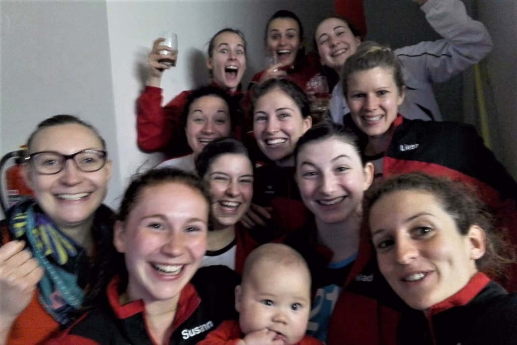 Gruppe lächelnder Frauen und eines Babys; viele tragen Sportoutfits und feiern gemeinsam in einem Raum. Eine hebt ein Glas.