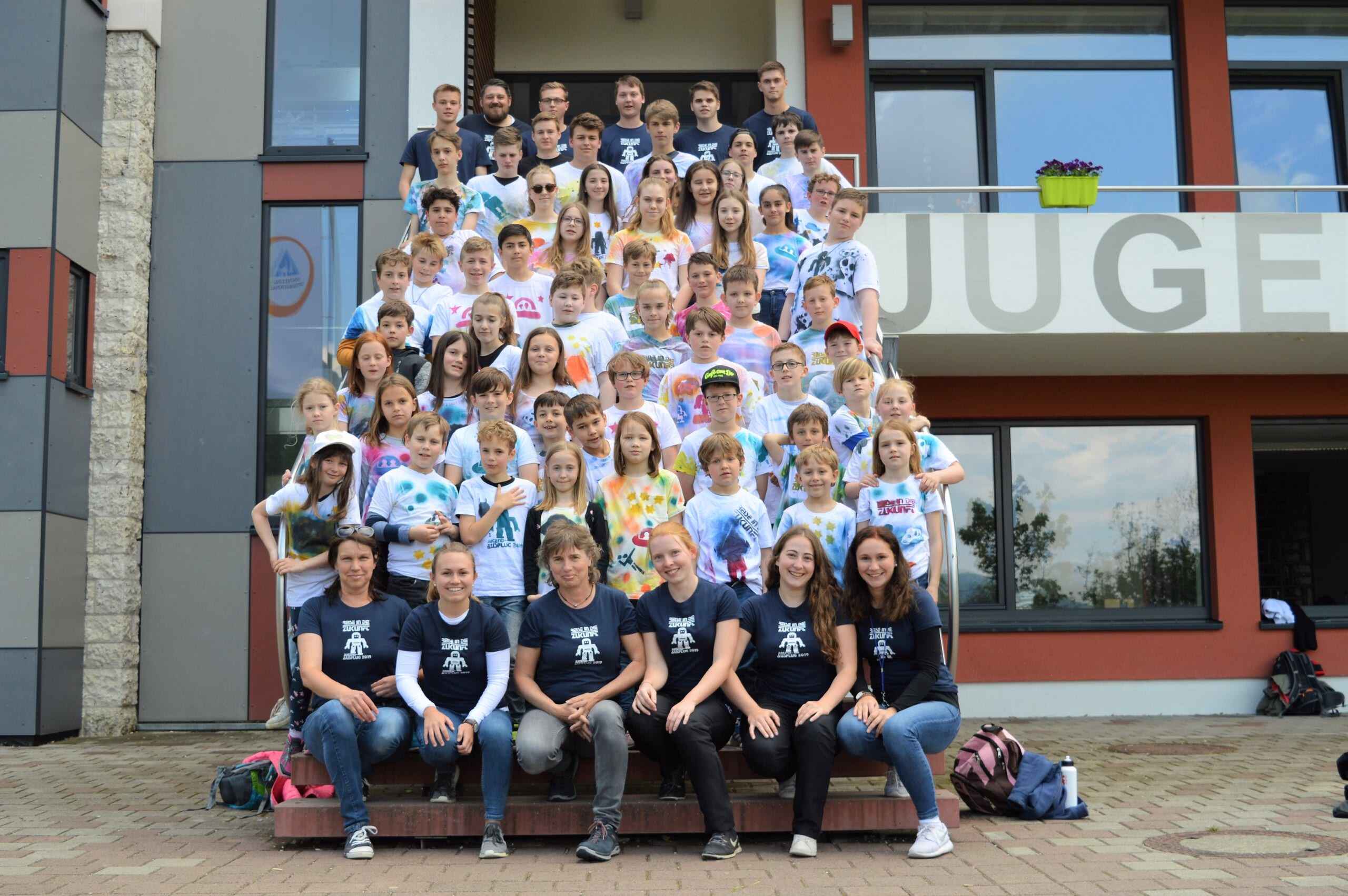 Gruppenfoto von Kindern und Betreuern vor einem Gebäudeingang, alle sitzen und stehen für einen gemeinsamen Jugend-/Ausflugsmoment.