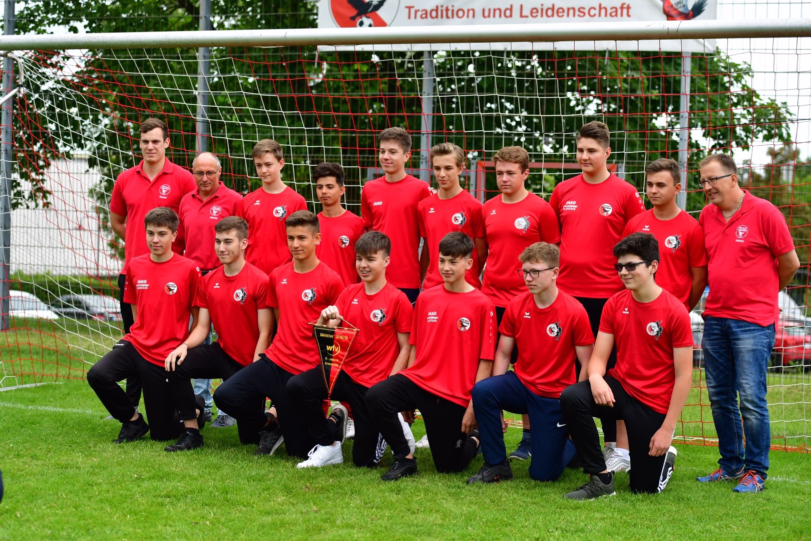 Mannschaftsbild mehrerer Jugendfußballer in roten Trikots vor einem Tor; einer hält einen Pokal, alle posieren auf dem Rasen.