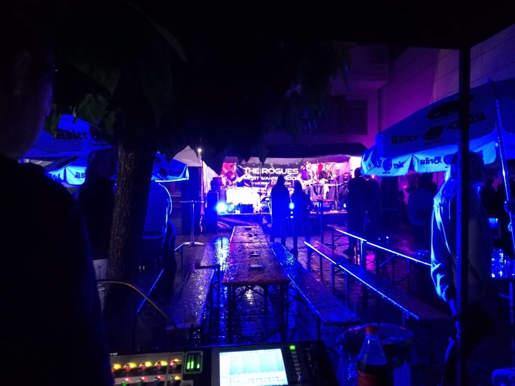 Danke! 7 Beleuchtete Bühne mit Musikern bei einem Straßenfest, Scheinwerfer in Blau und Violett mit Publikum im Vordergrund.