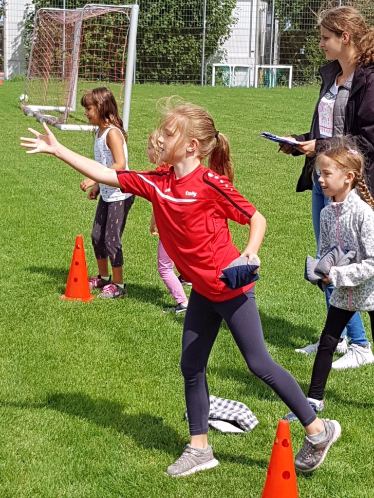 Ferienerlebnistag beim TSV Wiernsheim 6 Kinder üben Bewegung auf dem Rasen: eine Trainerin hält eine Sportübung an, Kinder balancieren und folgen mit Trainingshütchen.