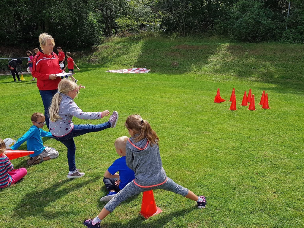 Ferienerlebnistag beim TSV Wiernsheim 7 Kinder im Freien bei einem Koordinationstraining: mehrere rote Kegel als Hindernisse, Anweisung durch eine Betreuungsperson.