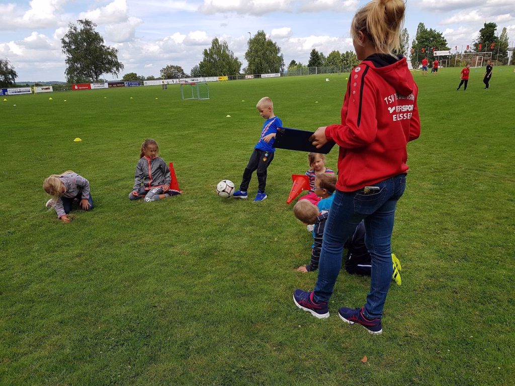 Ferienerlebnistag beim TSV Wiernsheim 8 Trainerin im roten Hoodie zeigt Kindern auf grünem Rasen mit Fußball und Markierungen, wie man im Spielraum agiert.