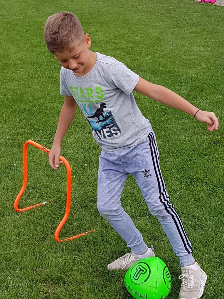 Ferienerlebnistag beim TSV Wiernsheim 9 Junge balanciert einen grünen Trainingsball auf dem Fußballrasen und bewegt sich dabei über ein orangefarbenes Hürden-Set.